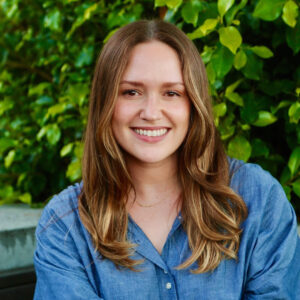 Photograph of Anna Balliet, Associate Marriage & Family Therapist. Anna is pictured outside, facing the camera and smiling. She is wearing a blue button down shirt. The background depicts green leaves.